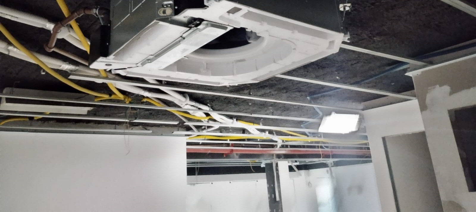 unidade evaporadora de ar-condicionado cassette sendo instalada em teto de obra, com fiação e tubulações apare