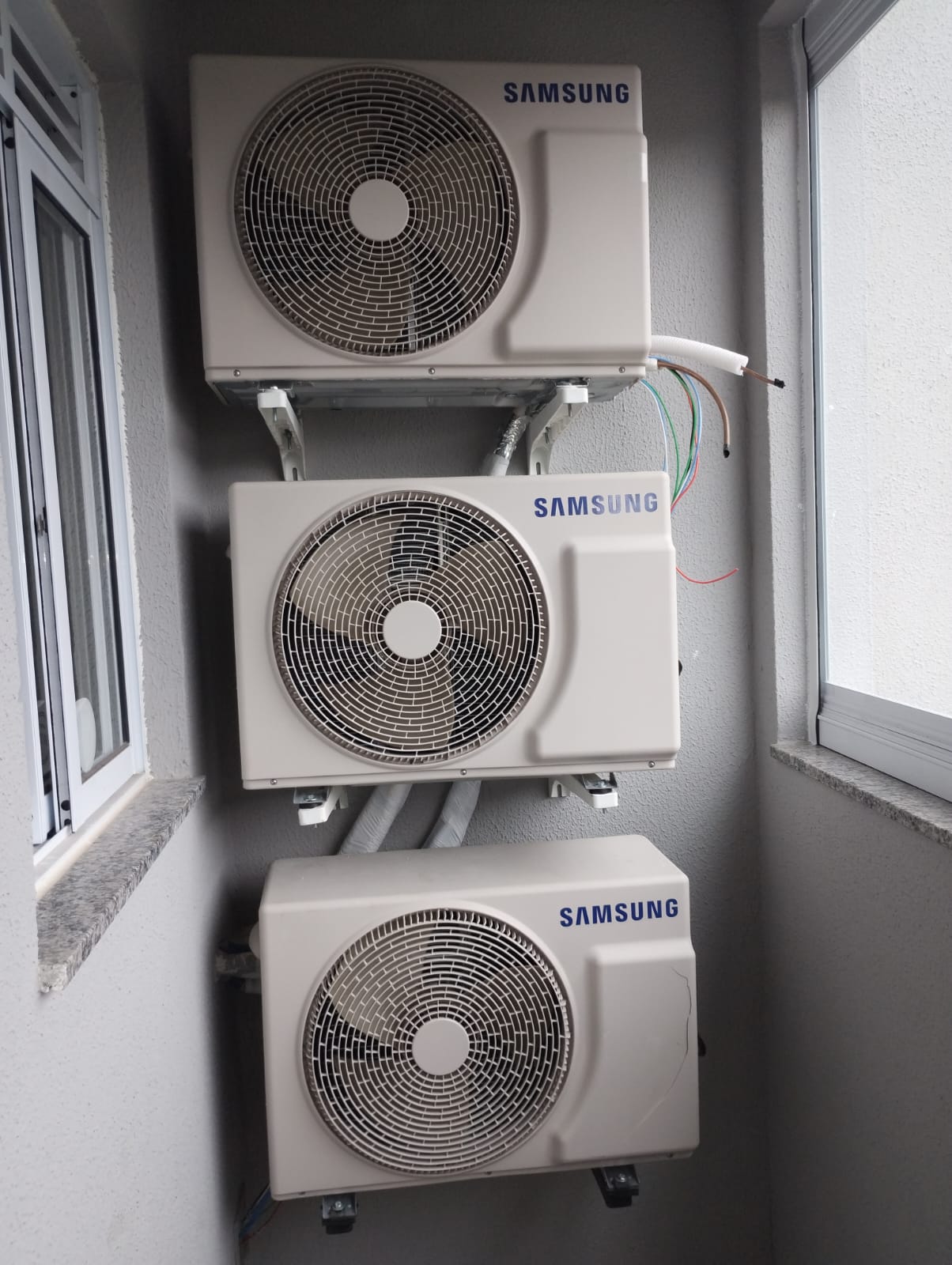 três condensadoras de ar-condicionado Samsung instaladas verticalmente numa parede de área externa entre janel